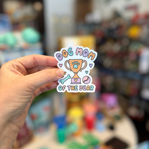 Hand holding a 'Dog Mom of the Year' sticker in front of a blurred store background