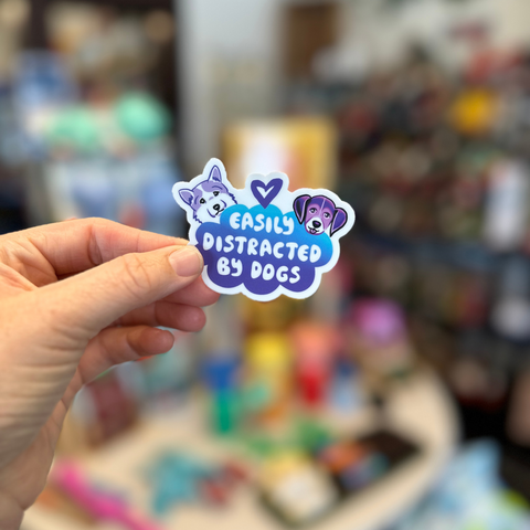Hand holding a sticker with 'Easily Distracted by Dogs' text and dog illustrations against a blurred background.