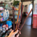 A hand holding four long, thin dog treats or sticks in a pet store, with a blurred background showing shelves and an indoor shop environment.