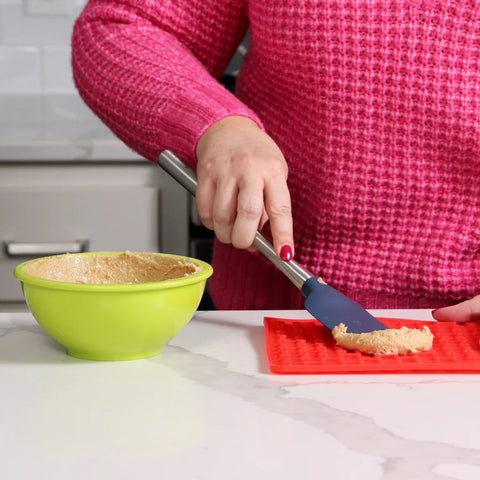 Person using a spatula to transfer batter from a green bowl onto a red silicone baking mat.