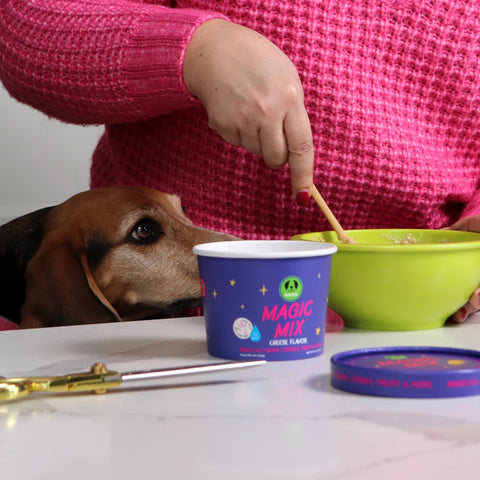Person feeding a dog from a 'Magic Mix' container with a green bowl and spoon on a table.