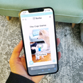 Person holding a smartphone displaying the Woofsie app with 'The Cup Game' on a carpeted floor.
